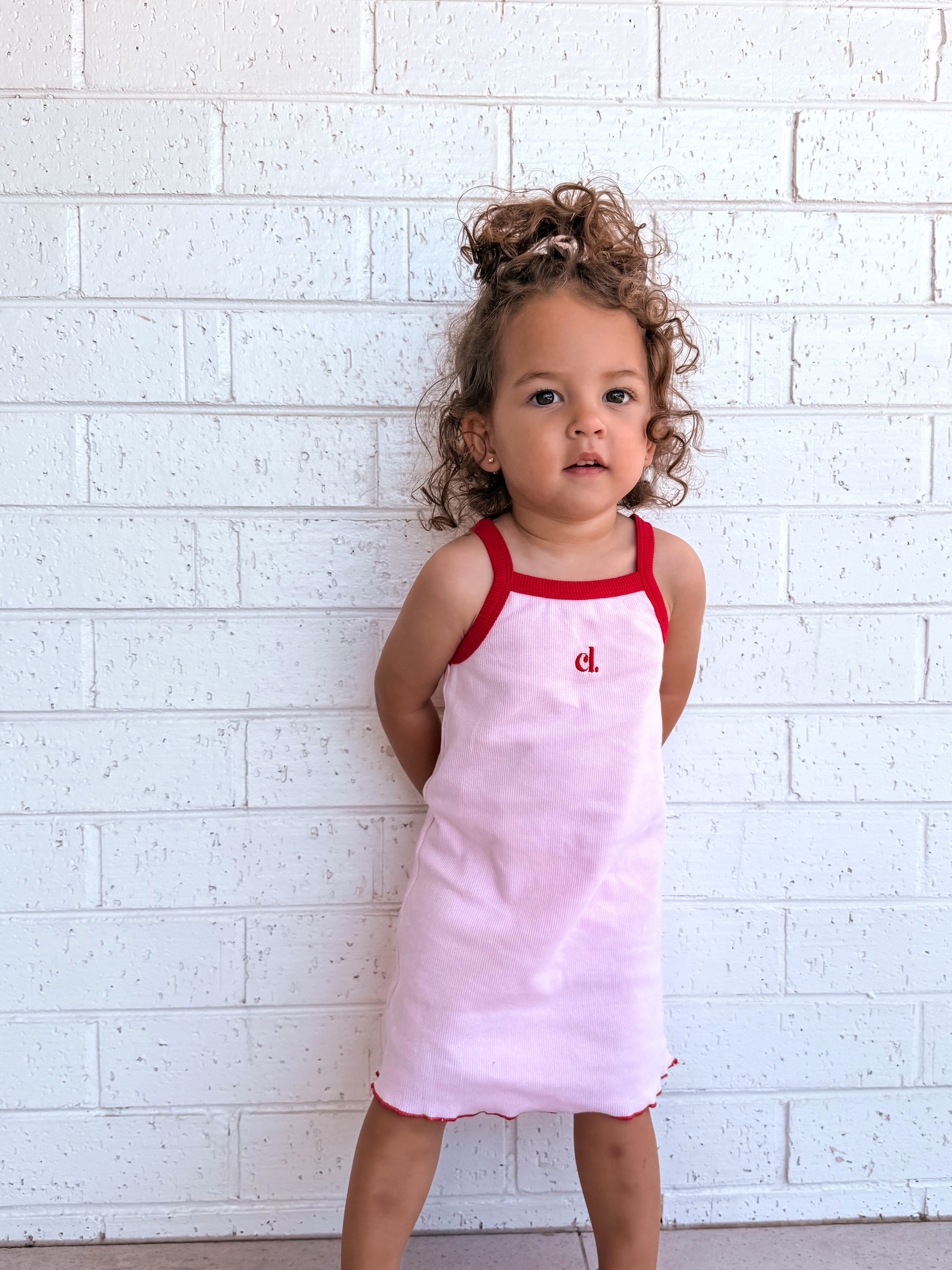 Child wearing a pink dress with red straps (Cami Lane Essential Ribbed Dress) against a white brick wall.